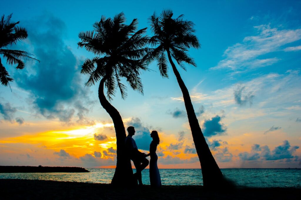 A couple holding hands against a palm-tree, with the ocean backdrop.