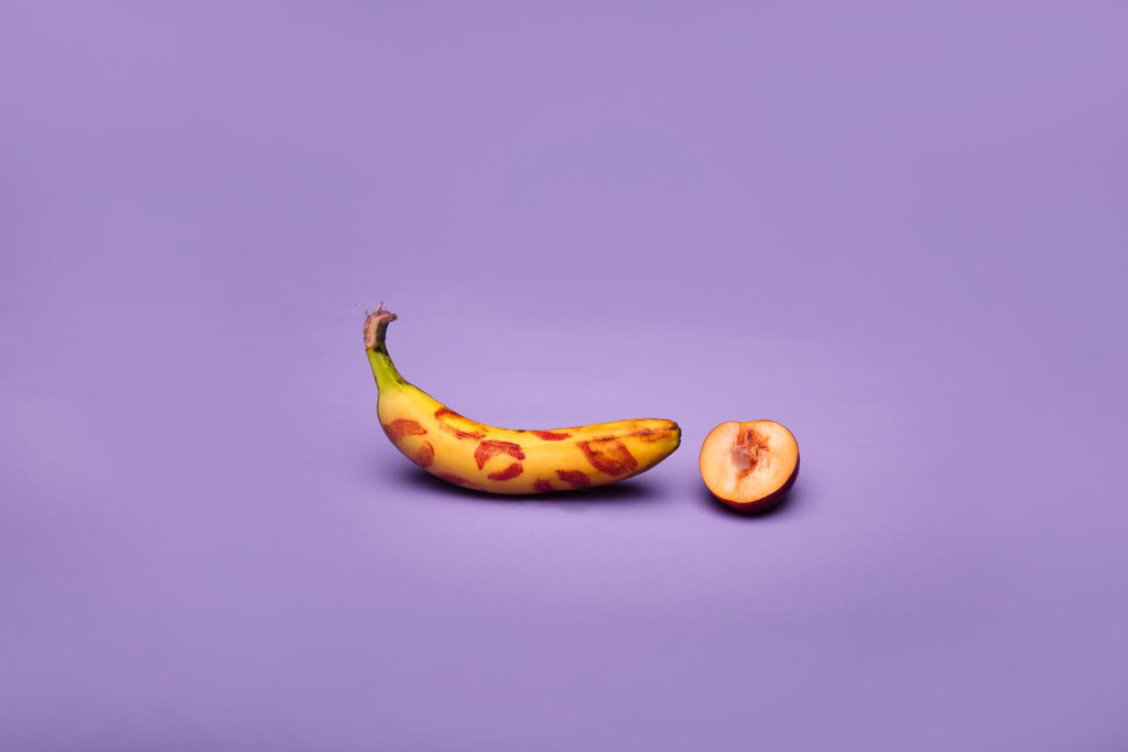 A banana with lipstick kisses all over it, sitting next to an open peach.